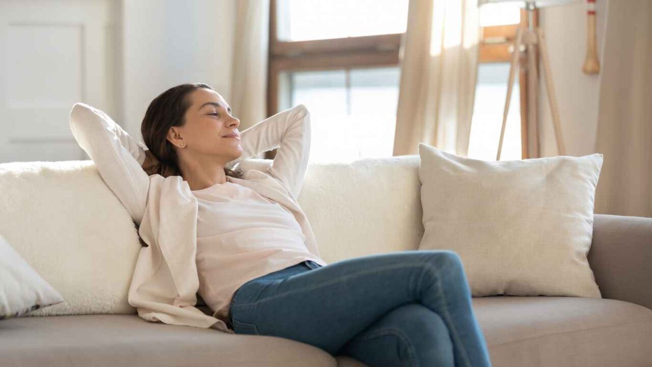 Carefree confident millennial female or teenage girl student enjoying moments of tranquility and comfort relaxing on sofa indoors switching on air conditioner and humidifier